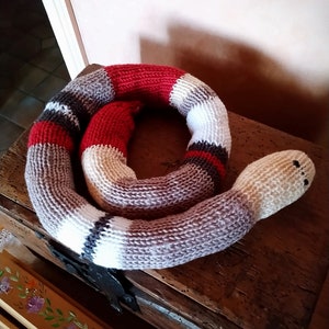 Puede incluir: Un juguete de serpiente de punto con un patrón de rayas rojas, blancas y grises. La serpiente está enroscada en un cofre de madera.