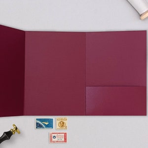 May include: A burgundy tri-fold invitation card with a pocket on the right side. The card is lying flat on a white surface with a few stamps and a wax seal nearby.