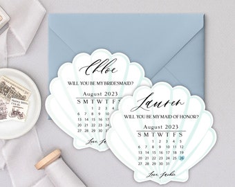 May include: Two light blue seashell-shaped cards with a calendar for the month of August 2023. The top card says "Chloe, Will you be my bridesmaid?" and the bottom card says "Lauren, Will you be my maid of honor?"