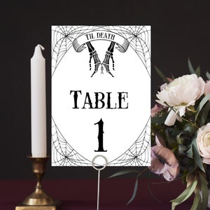 May include: A white table number card with black spiderweb design and the text "Til Death" above a pair of skeletal hands. The card reads "Table 1".
