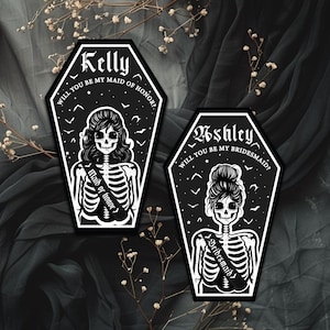 May include: Two black and white coffin-shaped cards with skeleton illustrations. One says "Kelly, Will you be my Maid of Honor?" and the other says "Ashley, Will you be my Bridesmaid?" Both feature decorative elements and a gothic theme.