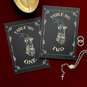 May include: Two rectangular black table number cards with gold foil lettering and illustrations. Each card features a hand holding a heart with the text TABLE NO. ONE and TABLE NO. TWO. The cards are decorated with gold filigree borders.