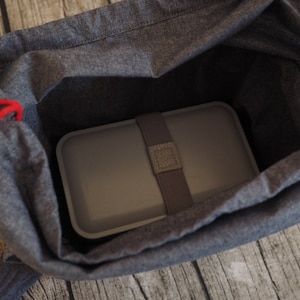 Op de afbeelding: Een grijze lunchtas met een rood trekkoord en een grijze lunchbox erin.