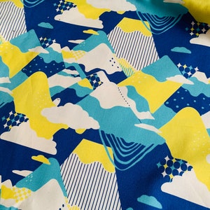 Op de afbeelding: Een blauwe stof met een herhalend patroon van gele, witte en blauwe geometrische vormen. De vormen lijken op bergen en wolken.