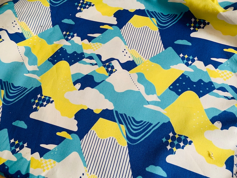 Op de afbeelding: Een blauwe stof met een herhalend patroon van gele, witte en blauwe geometrische vormen. De vormen lijken op bergen en wolken.