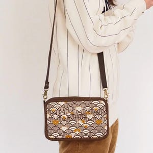 May include: A brown crossbody bag with a white and gold patterned flap. The pattern features a repeating design of waves and small animals.