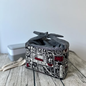 Op de afbeelding: Een grijze stoffen lunchtas met een zwart-witte Japanse print en rode accenten. De tas is vastgebonden met een grijze stoffen strik.