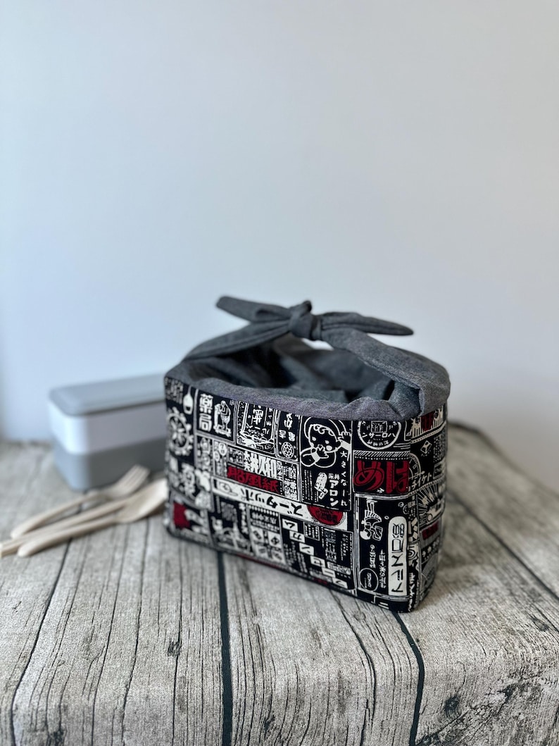 Op de afbeelding: Een grijze stoffen lunchtas met een zwart-witte Japanse print en rode accenten. De tas is vastgebonden met een grijze stoffen strik.