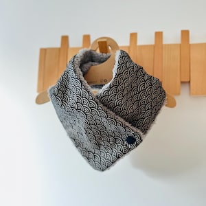 Puede incluir: Un calentador de cuello estampado en blanco y negro con forro de vellón gris. El calentador de cuello tiene un cierre de botón negro.