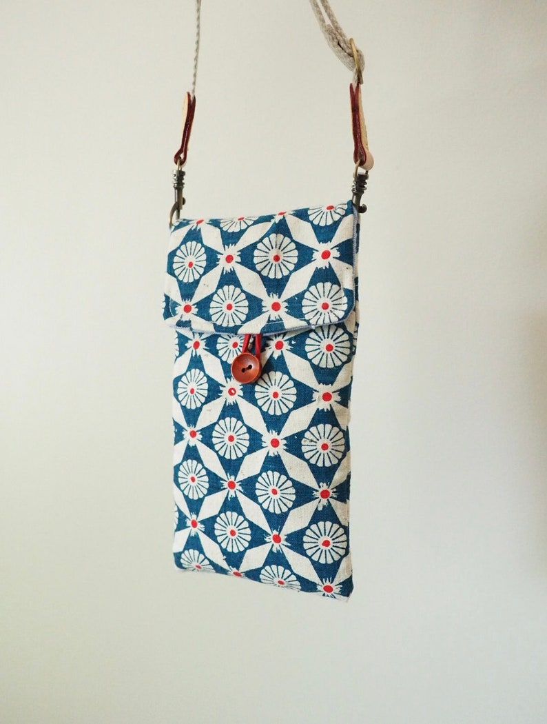 May include: A small blue and white patterned crossbody bag with a red button closure. The bag has a long strap and is made of fabric.