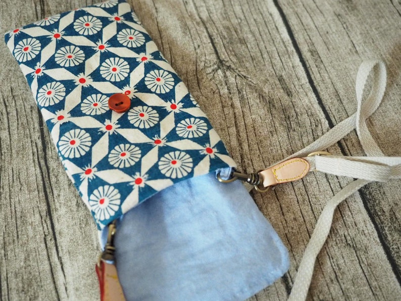 May include: A blue and white patterned fabric phone case with a red button and a beige strap. The case is open and the inside is lined with a light blue fabric.