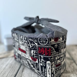 Op de afbeelding: Een grijze stoffen tas met een zwart-witte Japanse print met rode tekst. De tas heeft een geknoopte handvat.