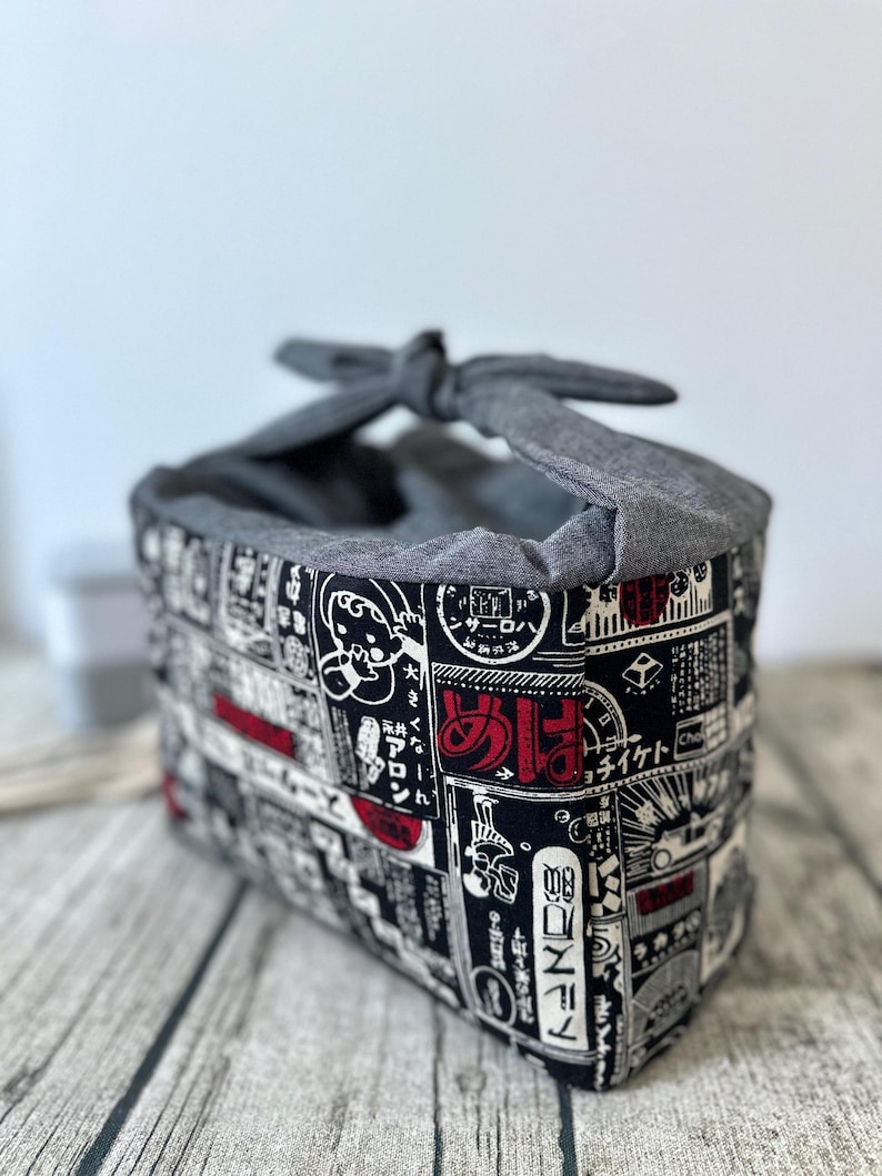 Op de afbeelding: Een grijze stoffen tas met een zwart-witte Japanse print met rode tekst. De tas heeft een geknoopte handvat.