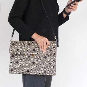 Puede incluir: Un bolso bandolera de patrón blanco y negro con una correa negra. El bolso tiene un cierre de solapa y un pequeño botón.