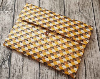 May include: A rectangular fabric sleeve featuring a repeating fan-shaped pattern in shades of mustard yellow, black, white, and tan. The sleeve has a fold-over flap secured by a single brown button and a thin elastic loop closure on the front center.