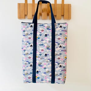 Op de afbeelding: Een rechthoekige draagtas met een herhaald patroon van katten, golven en geometrische vormen in roze, blauw en grijs. De tas heeft marineblauwe handvatten en afwerking. De tas hangt aan een houten kapstok.