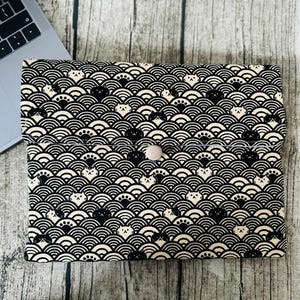 May include: A black and white laptop sleeve with a repeating pattern of cat faces and wave motifs. The sleeve is secured with a pale pink button. The sleeve is on a wooden surface next to a laptop.