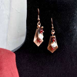 May include: Copper dangle earrings with a geometric design. Each earring features a copper-colored diamond shape, adorned with white and red accents. The earrings hang from copper-colored hooks.