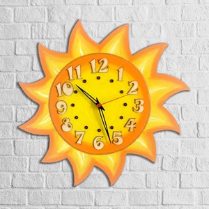 May include: A decorative wall clock shaped like a sun with a yellow and orange gradient design. The clock face is yellow with wooden numerals and black hands. The clock is mounted on a white brick wall.