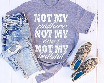 May include: A heather gray t-shirt with the text "NOT MY pasture NOT MY cows NOT MY bullshit" in white. The shirt is paired with light wash denim jeans and camouflage print sneakers.