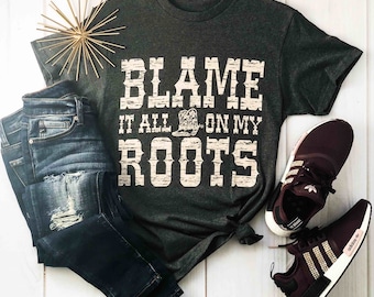 May include: A charcoal grey t-shirt with the slogan "BLAME IT ALL ON MY ROOTS" in a distressed font, alongside blue denim jeans and burgundy trainers. A gold starburst ornament adds a decorative touch, creating a casual, country-inspired aesthetic.