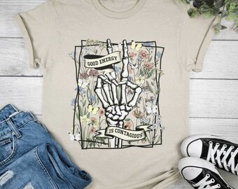 May include: Beige t-shirt featuring a skeleton hand making a peace sign, surrounded by a floral design with butterflies. The shirt has the text "GOOD ENERGY IS CONTAGIOUS" on banners. The shirt is paired with blue jeans and black canvas sneakers.