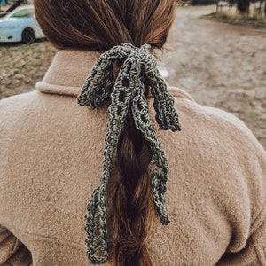 May include: A gray crocheted hair tie is tied around a brown braid. The hair tie is made of a delicate, open-weave pattern. The person is wearing a tan coat. The background is a blurred outdoor scene with trees and a car.