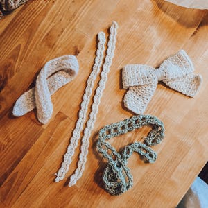 Könnte beinhalten: Vier gehäkelte Gegenstände auf einer Holzoberfläche. Zwei weiße Häkelborten, eine weiße HäkelSchleife und eine grüne Häkelborte.