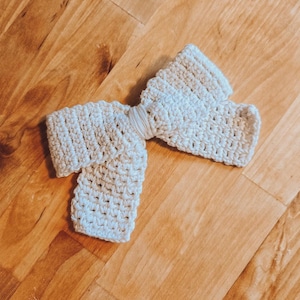 Könnte beinhalten: Eine gehäkelte Schleife aus hellbeigem Garn. Die Schleife ist verknotet und hat ein schlichtes, elegantes Design.