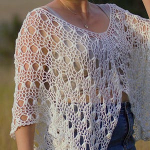 May include: White crochet poncho with a delicate openwork pattern. The poncho has a loose, flowing fit and is perfect for layering over a tank top or t-shirt.