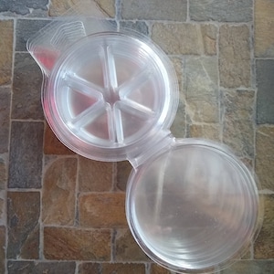 May include: A clear, plastic container with a hinged lid, open to reveal multiple clear, round, shallow trays. The container is designed with a central, star-shaped divider. The trays are stacked and ready for use.