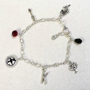 May include: Silver charm bracelet with multiple charms: a red gem, an arrow, a rose, a skull, a black gem, a tree, the letter "K", and a circular emblem. The bracelet is made of linked chain.