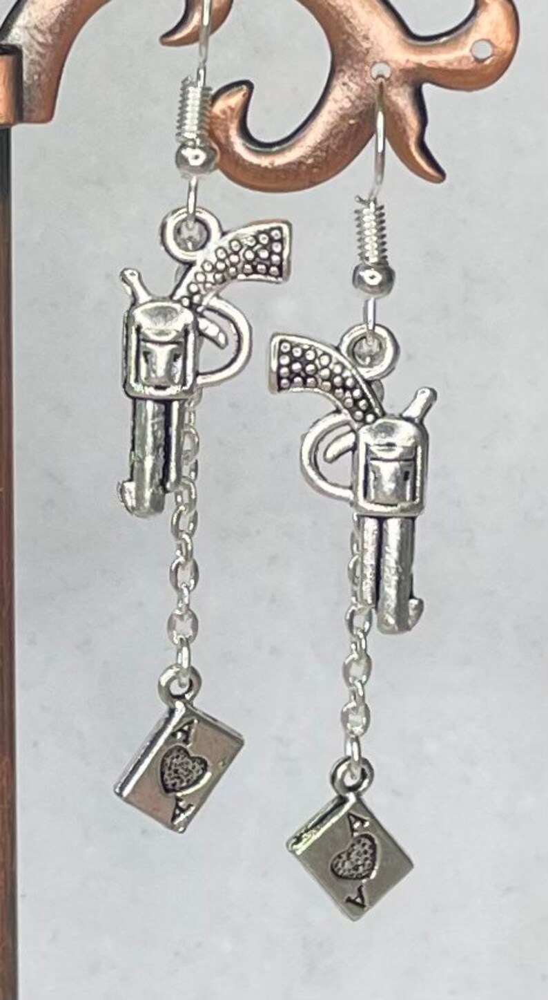 Puede incluir: Pendientes de tono plateado con un peque&ntilde;o dije de pistola y un dije de as de corazones colgante.