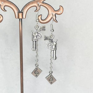 Puede incluir: Pendientes de tono plateado con un dije de pistola y un as de corazones colgando de una cadena plateada.