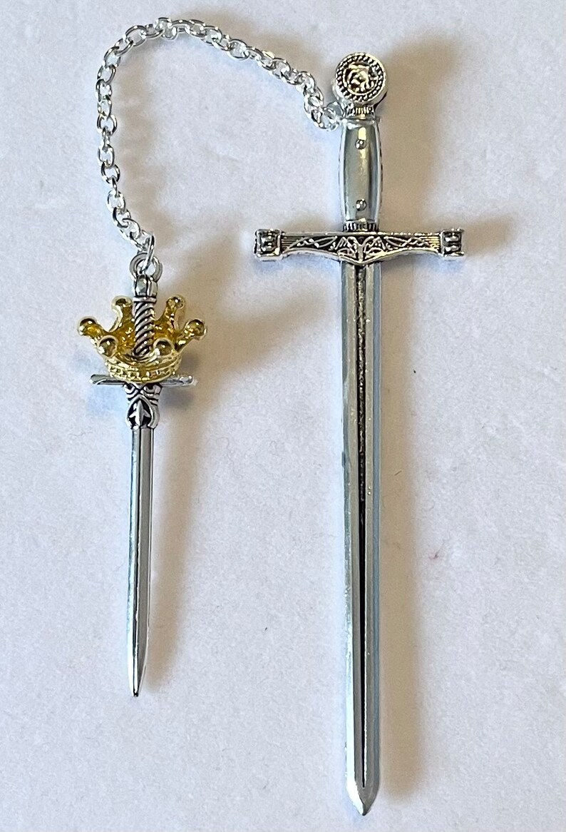 May include: Two silver-toned swords with gold accents. The smaller sword has a crown on the hilt and is attached to the larger sword by a silver chain.