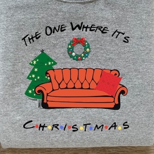 May include: Grey sweatshirt with the text "The One Where It's Christmas" in black. The text is surrounded by a Christmas wreath, a Christmas tree, and a red couch with a red pillow that says "I'll be there for you".