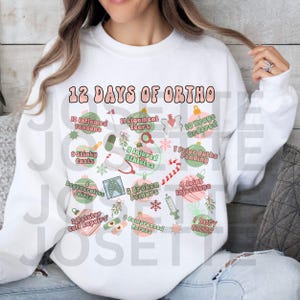 May include: White sweatshirt with the text "12 Days of Ortho" in pink and green. The design includes illustrations and text related to orthopedic conditions and treatments, such as "Inflamed Tendons" and "Broken Femurs."