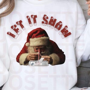 Puede incluir: Sudadera blanca con una ilustración de Papá Noel rojo y blanco y el texto "Let It Snow" en letras rojas. Papá Noel sostiene una pajita y una sustancia blanca en una bandeja plateada.