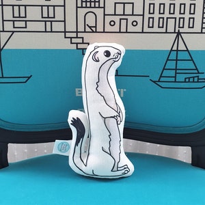 May include: White plush toy shaped like a weasel or stoat. The toy is standing upright with its paws together. The toy has black eyes and a black nose. The toy is sitting on a blue chair with a black frame.