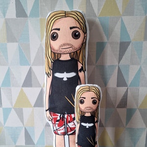 May include: Two plush dolls, one larger than the other, are both dressed in black tank tops with a white bird design, red and black patterned shorts, and black sneakers. Both dolls have long blonde hair and large brown eyes.