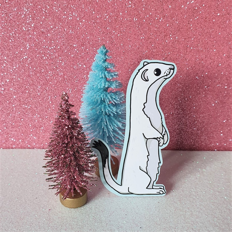 May include: A white weasel sticker with a black-tipped tail, standing upright. The sticker has a light blue border. Two small decorative Christmas trees are behind the weasel, one pink and one blue, against a pink glitter background.