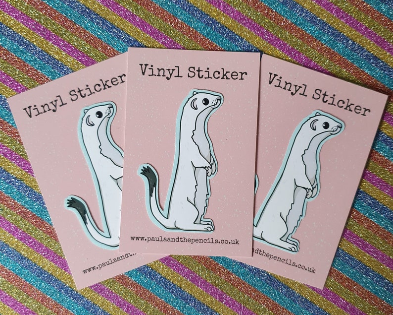 May include: Three vinyl stickers featuring a cartoon ermine standing upright on a pink background. The stickers have a white body, black tail tip, and are outlined in light blue. Each card reads "Vinyl Sticker" and includes a website address.