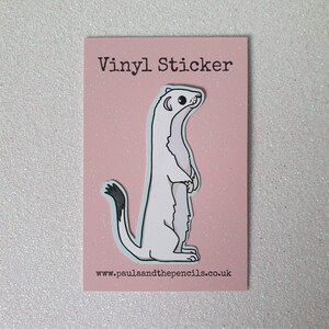 May include: A vinyl sticker featuring a cartoon ermine standing upright on a pink background. The ermine is white with a black-tipped tail and a light blue outline. The text "Vinyl Sticker" is at the top, and the website address is at the bottom.