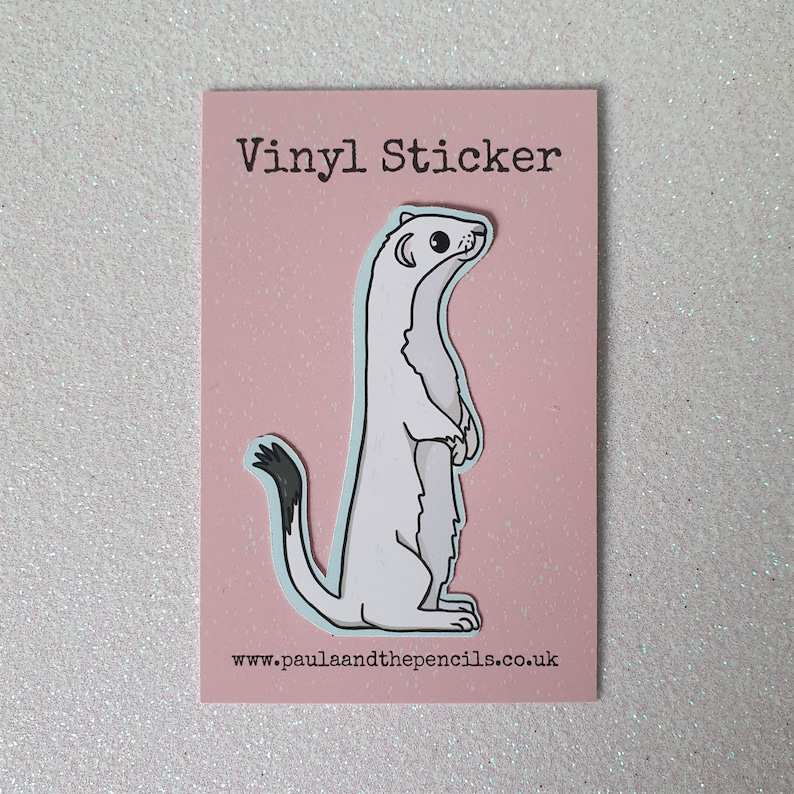 May include: A vinyl sticker featuring a cartoon ermine standing upright on a pink background. The ermine is white with a black-tipped tail and a light blue outline. The text "Vinyl Sticker" is at the top, and the website address is at the bottom.