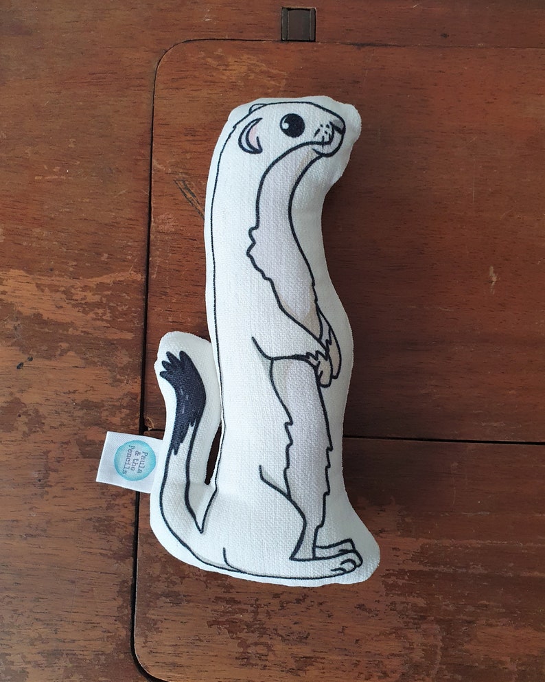 May include: White plush toy shaped like a weasel with black eyes and a black tail. The toy is standing upright and has a tag that says "Pilla & the Priscilla".