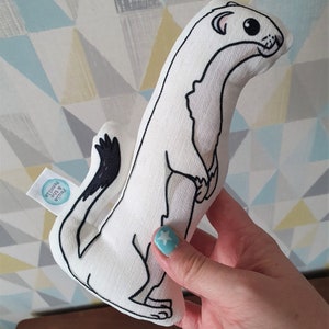 May include: White plush toy shaped like a weasel with black eyes and a black tail. The toy is held in a hand with blue nail polish. The toy has a small tag that says "Petite & Precious".