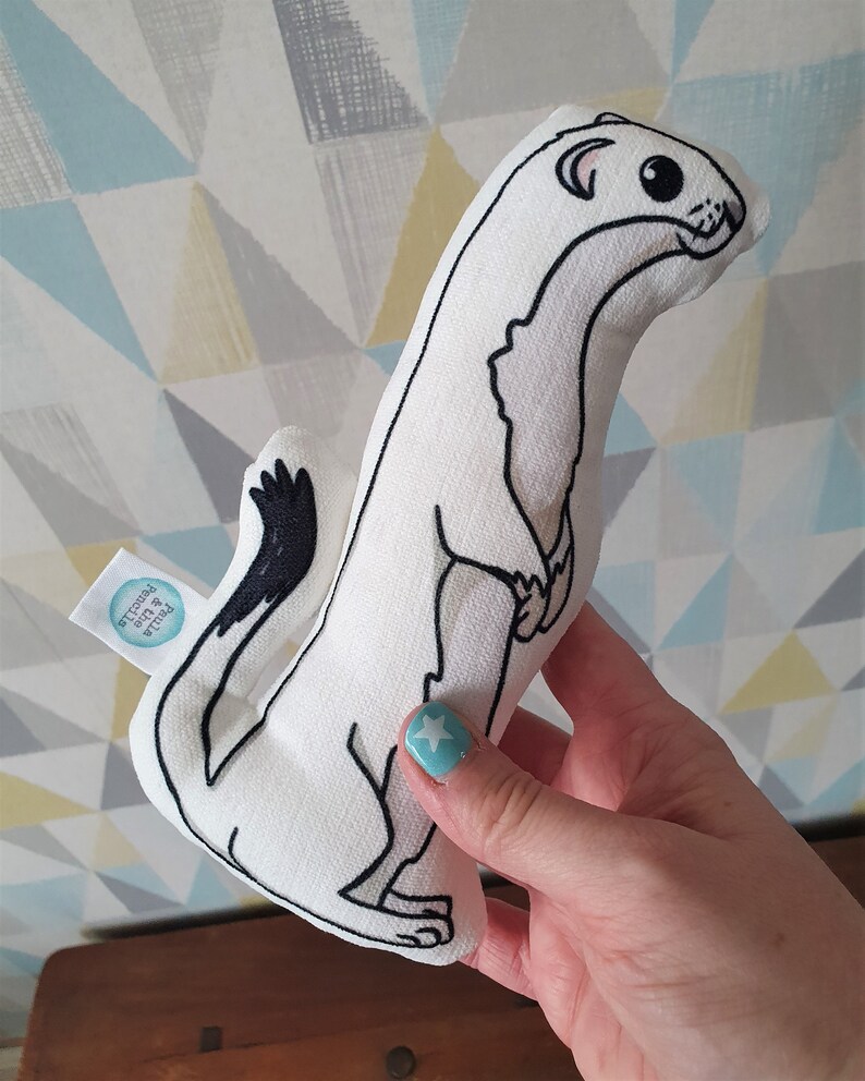 May include: White plush toy shaped like a weasel with black eyes and a black tail. The toy is held in a hand with blue nail polish. The toy has a small tag that says "Petite & Precious".