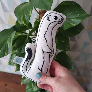 May include: White plush toy shaped like a weasel with black eyes and a black tail. The toy is being held in a hand with a blue nail polish.