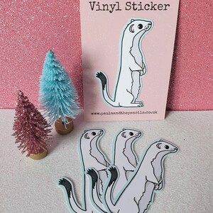 May include: A collection of vinyl stickers featuring a cartoon weasel design. The weasels are white with black tail tips and stand upright. The stickers are on a light blue background. A pink card with the text "VINYL STICKER" is also visible.