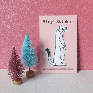 May include: A white vinyl sticker of a weasel standing on its hind legs with its tail curled around its body. The sticker is on a pink background with the text "Vinyl Sticker" at the top and "www.paulaandthepencils.co.uk" at the bottom.
